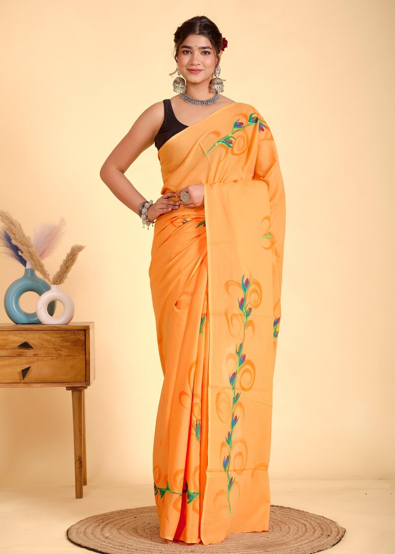 May include: A woman wearing an orange saree with a floral design in green, blue, and orange. The saree is draped over the woman's body. She is wearing a black top and silver jewelry. The background is a light orange color.