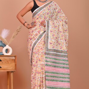 May include: A white saree with a floral pattern in yellow, pink, and green. The border features stripes and floral designs. The saree is worn over a black top.