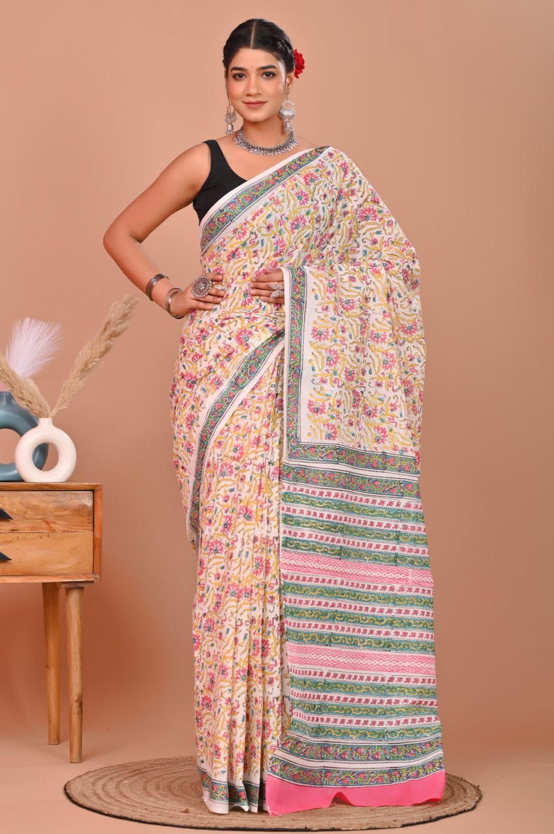 May include: A white saree with a floral pattern in yellow, pink, and green. The border features stripes and floral designs. The saree is worn over a black top.