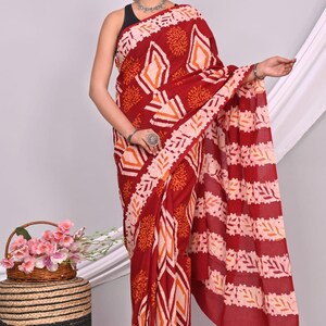 May include: A maroon saree with a geometric and floral pattern in shades of orange and white. The saree is draped over a person, showcasing the intricate design. The background is a neutral white.