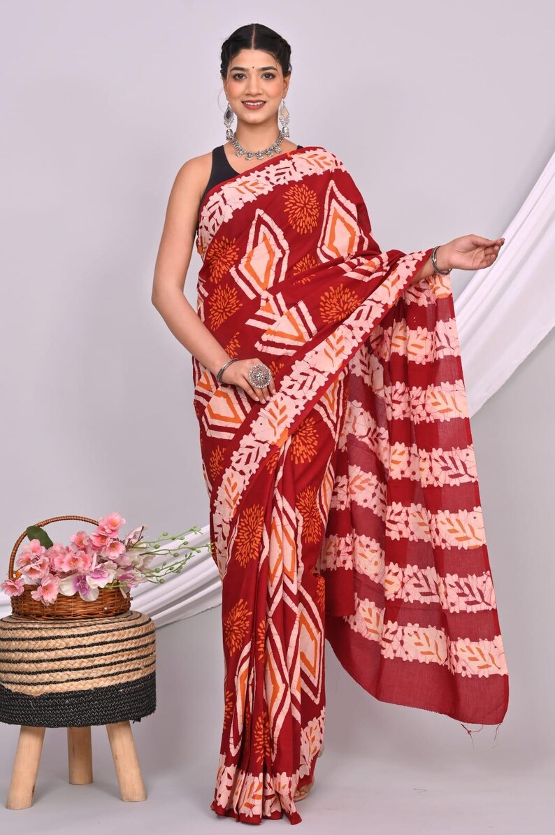 May include: A maroon saree with a geometric and floral pattern in shades of orange and white. The saree is draped over a person, showcasing the intricate design. The background is a neutral white.