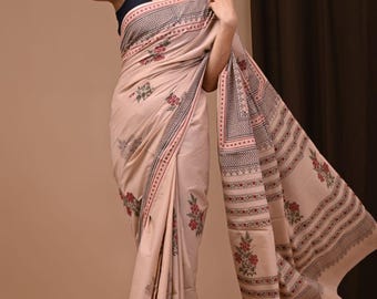 May include: A light peach-colored saree with a floral pattern and decorative border. The saree features a repeating design of red flowers and geometric shapes. The model is wearing a black top and silver jewelry.