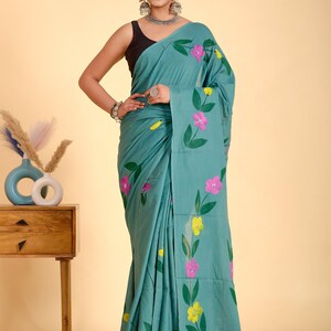 May include: Teal saree with a floral pattern in pink and yellow. The flowers are hand-painted with green leaves. The saree is draped over a person. The background is a light beige color.