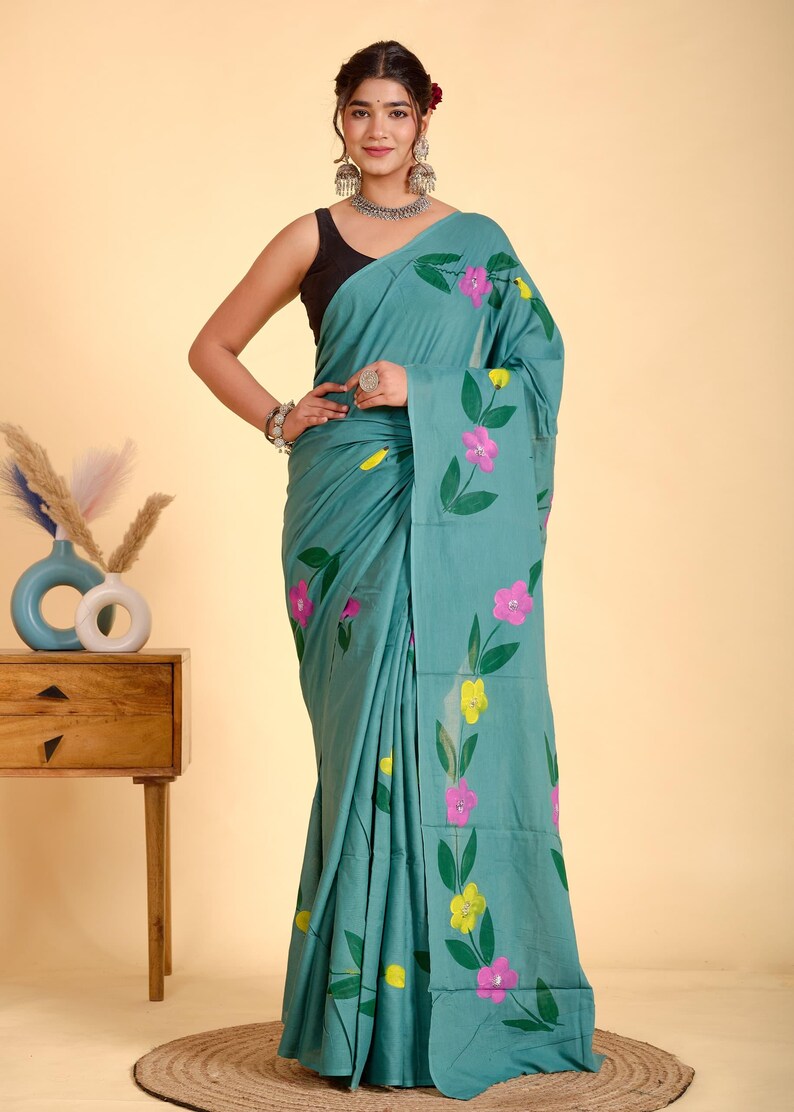 May include: Teal saree with a floral pattern in pink and yellow. The flowers are hand-painted with green leaves. The saree is draped over a person. The background is a light beige color.