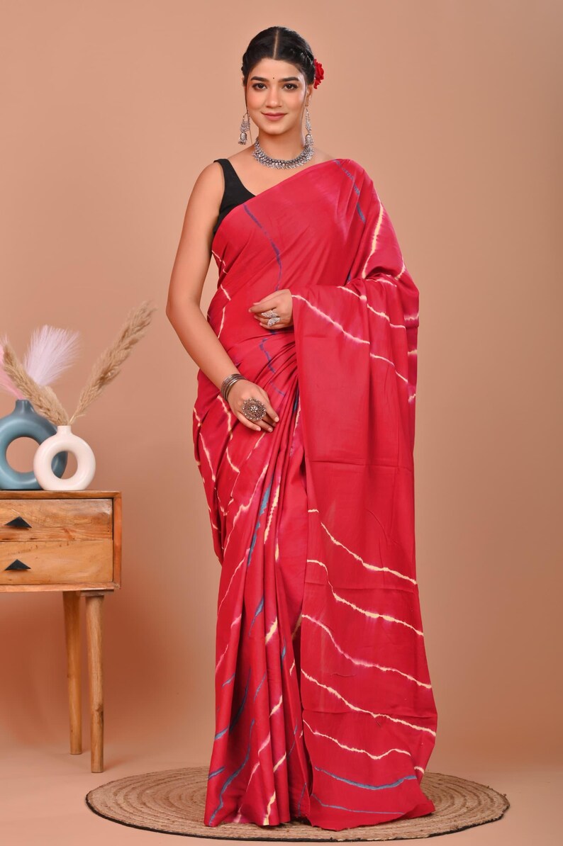May include: A vibrant red saree with white and blue striped patterns. The traditional Indian garment is draped over a person, showcasing the intricate design and flowing fabric. The saree is a bold statement piece.