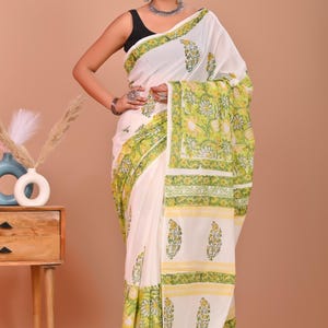 May include: White saree with a floral pattern in shades of green and yellow. The saree features a decorative border and is draped over a person. The background is a solid peach color.