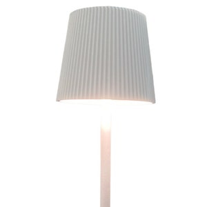 May include: A white table lamp with a square base and a tall, slender stem. The lampshade is a light, textured white, and the lamp is illuminated. The lamp is a modern design, suitable for a living room or bedroom.