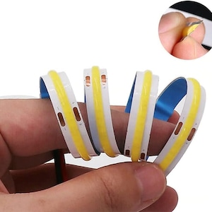 May include: Flexible LED light strips with a bright yellow center and white edges. The strips are shown coiled, with a blue backing visible. A close-up shows the adhesive backing being peeled. Wires are attached to the end of the strip.