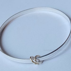 Könnte beinhalten: Ein silbernes Armreif-Armband mit einem kleinen goldenen Ring-Anhänger. Das Armband ist schlicht und elegant.