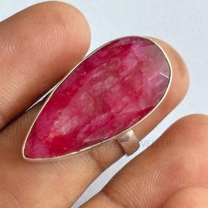May include: A large, teardrop-shaped ring featuring a vibrant, faceted ruby gemstone. The ruby is set in a silver band, showcasing its deep red color. The ring's design is simple, highlighting the beauty of the stone.