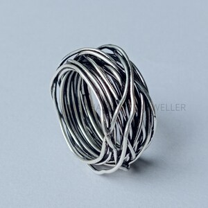 Può includere: Un anello color argento con un design intrecciato. L'anello è realizzato con più fili di metallo intrecciati per creare un aspetto strutturato, simile a un nido. L'anello è mostrato su uno sfondo grigio chiaro.