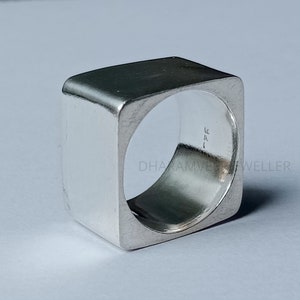 May include: A square, silver ring with a hollow interior. The ring has a smooth, polished surface and a modern, geometric design. The ring is a bold statement piece of jewelry.