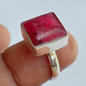 May include: A silver ring with a large, square-cut, deep red gemstone. The gemstone is set in a silver bezel, and the ring band is smooth and polished. The ring is a statement piece, highlighting the vibrant colour of the stone.