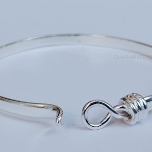 Puede incluir: Un brazalete de plata con un diseño de nudo retorcido. El brazalete está abierto y tiene un estilo simple y elegante.
