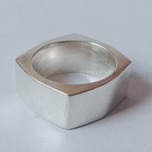 Puede incluir: Un anillo cuadrado de plata con un acabado liso y pulido. El anillo es grueso y tiene una banda ancha.
