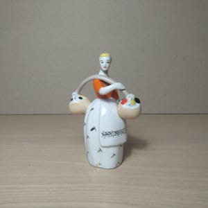 Könnte beinhalten: Keramikfigur einer Frau, die zwei Körbe trägt. Die Frau trägt ein orangefarbenes Oberteil und ein weißes Kleid mit schwarzen und blauen Akzenten. Die Körbe sind beige mit farbenfrohen Details.