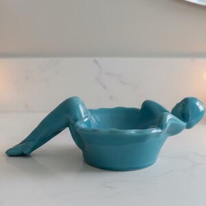 May include: A teal ceramic bowl in the shape of a reclining figure. The bowl has a rounded base and a scalloped rim. The figure's legs extend outward, and its arms are bent, supporting the bowl. The bowl is on a white surface.