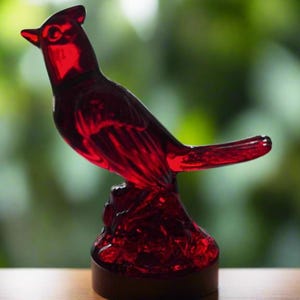 Könnte beinhalten: Eine rote Glasfigur eines Kardinals mit detailliertem Design. Die Figur steht auf einem Sockel mit strukturierter Oberfläche.