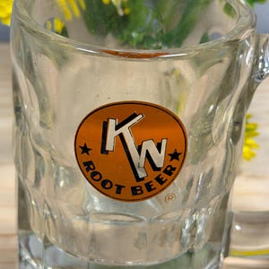 May include: Clear glass root beer mug with a handle. The mug features a circular orange and brown label with the text "KW ROOT BEER" and two stars. The mug is empty and has a textured exterior.