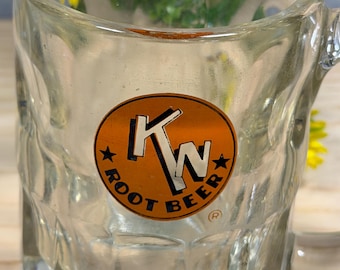 May include: Clear glass root beer mug with a handle. The mug features a circular orange and brown label with the text "KW ROOT BEER" and two stars. The mug is empty and has a textured exterior.