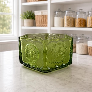 May include: A square, green glass bowl with embossed star and eagle designs. The bowl is sitting on a white countertop in a kitchen setting. Shelves in the background hold jars of food and woven baskets.