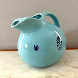 Può includere: Una brocca in ceramica azzurra con corpo arrotondato, manico e beccuccio. La brocca presenta due disegni floreali blu scuro. La brocca viene probabilmente utilizzata per servire bevande.
