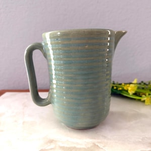 May include: A handmade ceramic pitcher in shades of blue and gray. The pitcher has a cylindrical shape with a handle and a spout. Horizontal ridges encircle the body, adding texture. The pitcher sits on a white surface.