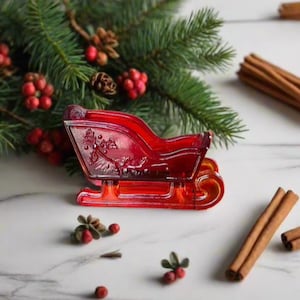 Może przedstawiać: Sanki bożonarodzeniowe z czerwonego szkła z wzorem ostrokrzewu. Sanki stoją na białym marmurowym blacie, wokół nich rozrzucone są czerwone jagody i cynamonowe patyczki.
