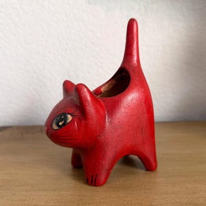 May include: A red cat-shaped planter with a tall, upright tail. The ceramic cat has a small opening on its back for planting. The cat's face has painted details, including eyes and whiskers. The planter is on a wooden surface.