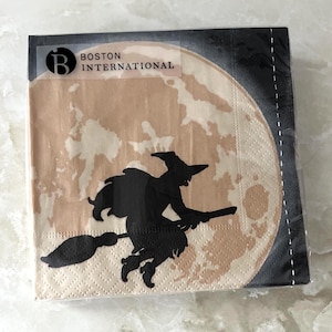 Puede incluir: Un paquete de servilletas de papel con temática de Halloween. Las servilletas presentan una silueta negra de una bruja volando en una escoba frente a una luna llena. Se ve la marca "BOSTON INTERNATIONAL".