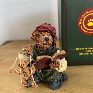May include: A Boyd's Bears figurine depicts a brown bear wearing a red hat and reading a book. The bear holds a small teddy bear on a swing. The figurine is detailed with glasses and a green tunic. A green box with gold lettering is in the background.
