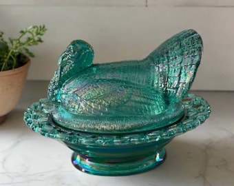 May include: A teal blue glass turkey butter dish with a detailed feathered design. The dish is sitting on a white marble surface.