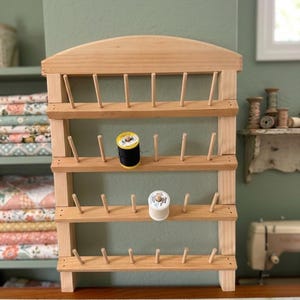 May include: A wooden thread rack with multiple pegs for holding spools of thread. The rack is made of light-colored wood and has three shelves. Two spools of thread, one black and one white, are visible on the rack. The background is a light green wall.
