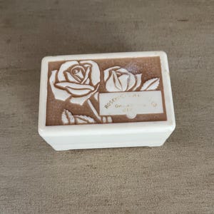 May include: A rectangular, off-white jewelry box with a brown lid. The lid features a relief design of two roses and the text "Rosefield Jewelers, Garden City". The box is closed and sits on a neutral surface.