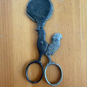 May include: A metal tool shaped like a rooster with a circular blade at the top and two finger loops at the bottom. The rooster figure features detailed engravings on its body, a small screw at the center, and a dark, weathered finish throughout.