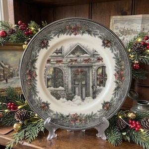 May include: A round ceramic plate featuring a central illustration of a brick building decorated for the holidays with snow, wreaths, and red bows. The rim is dark gray with a repeating pattern of pine branches, pinecones, and red ribbons.