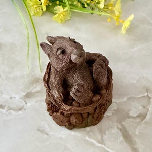 May include: A brown resin sculpture of a rabbit nestled in a textured, round basket. The rabbit has detailed fur and is posed with its head up. The basket is also brown and has a rough, woven appearance. Yellow flowers and green stems are in the background.