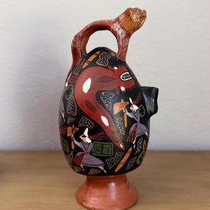 May include: A black ceramic vessel with a terracotta-colored base and handle shaped like an animal head. The body features hand-painted illustrations of figures in purple and white clothing, a large red creature, and geometric shapes in orange and green.