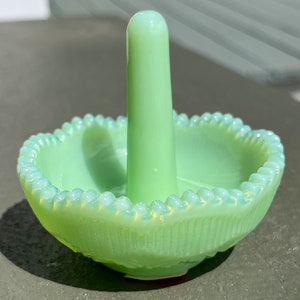May include: A light green glass ring holder with a central post and a scalloped rim. The dish has a textured pattern on the sides and a smooth, rounded base. The ring holder is approximately 4 inches tall.