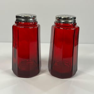 May include: Two red glass salt and pepper shakers with silver-colored metal lids. The shakers have a faceted, rectangular shape. The lids have small holes for dispensing the contents.