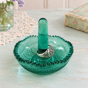 May include: A teal green glass ring holder featuring a central vertical post and a circular base with a scalloped edge and embossed floral patterns. A silver-toned metal ring with an intricate openwork filigree design rests on the central post.
