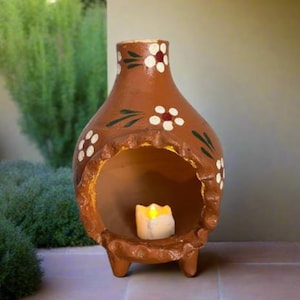 Puede incluir: Un chimenea de color terracota con una vela dentro. La chimenea tiene un cuerpo redondeado, una parte superior acampanada y tres patas pequeñas. Flores blancas con centros rojos y hojas verdes decoran el exterior. La vela emite un brillo amarillo cálido.