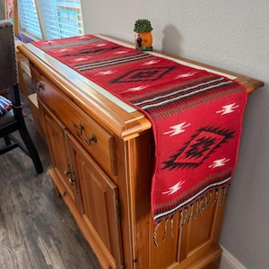May include: A red table runner in a Southwestern style, featuring a geometric pattern in black, white, and brown. The runner, with fringed ends, is displayed on a wooden cabinet.