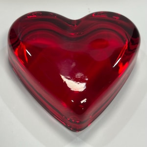 May include: A heart-shaped, red glass object. The glass is a deep, translucent red with darker red streaks. The object is smooth and polished, reflecting light. The shape is a classic heart, with rounded curves and a flat base.