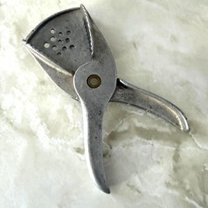 May include: A vintage, silver-toned citrus juicer. The juicer has a hinged design with a perforated bowl for catching seeds and pulp. The handle is curved for easy gripping. The juicer is made of metal and appears to be well-used.