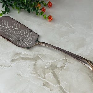 May include: Silver-plated cake server with an ornate, engraved design on the blade. The server features a long, curved handle and a decorative edge. The blade is rectangular with a scalloped edge and a detailed, symmetrical pattern.