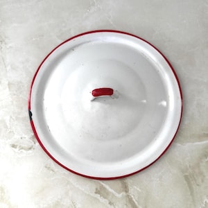 May include: White enamel pot lid with a red rim and a small red handle. The lid is round and appears to be vintage or retro in style. The lid is on a marble surface.