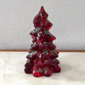 May include: A decorative red glass Christmas tree with green leaf accents. The tree is a vibrant shade of red and has a glossy finish. The tree is a festive holiday decoration.