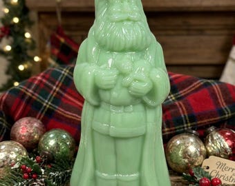 May include: A pale green, translucent glass figurine of Santa Claus standing in a long coat with a beard and hat. The figure is molded with detailed facial features, a belt, and hands held together at the chest, showcasing a smooth, glossy finish.