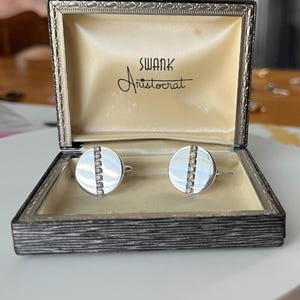 Puede incluir: Gemelos plateados con una fila de pequeñas piedras transparentes en el centro de cada cara circular. Los gemelos se exhiben en una caja negra con las palabras "SWANK Aristocrat" impresas en el interior.